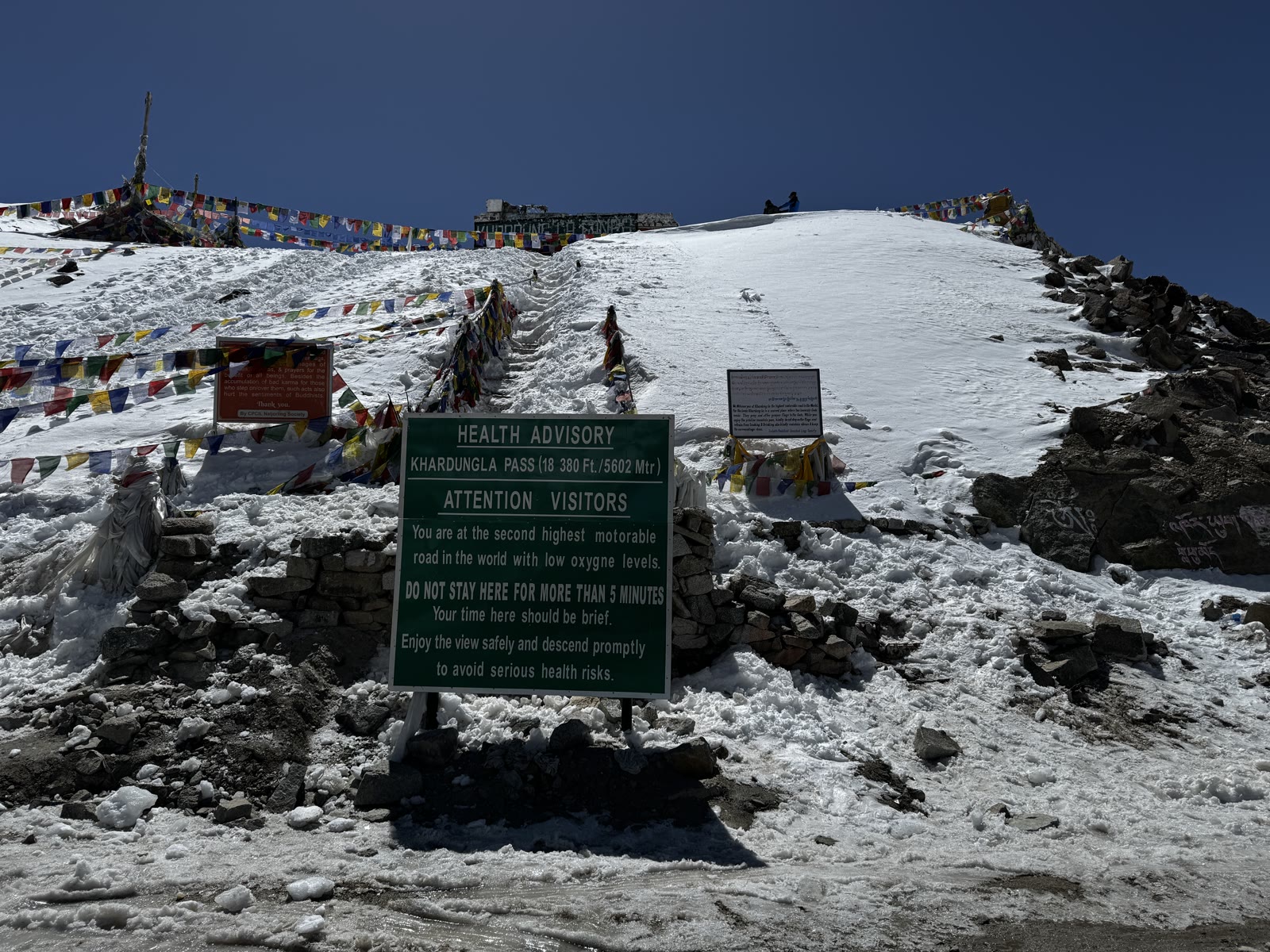 Health advisory board at Khardung La Pass (18,380 ft / 5602 m) — prayer flags and snow-covered peaks behind